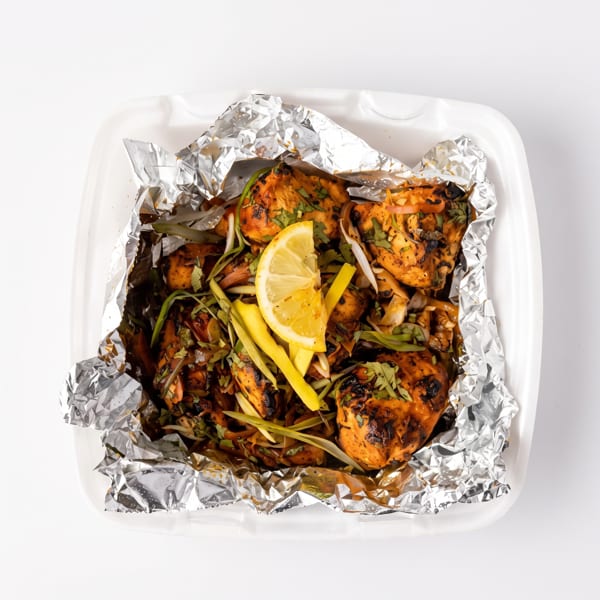 Grilled chicken pieces with herbs, onions, and a lemon wedge in a foil-lined takeout container.